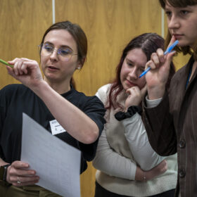 Zbliżenie na 3 młode dziewczyny stojące w pomieszczeniu. Dziewczyny przyglądają się czemuś co jest poza kadrem. Dziewczyna stojąca najbardziej po lewej stronie ma rude włosy. Ma też wyciągniętą lewą rękę, w której trzyma zielony długopis. Tłumaczy coś pozostałej dwójce. Dziewczyna w środku pochyla się do przodu. Dłonią podpiera głowę. Trzecia z dziewczyn stoi najbardziej po prawej. Trzyma w ręce niebieski długopis. Przykłada go do ust w zamyśleniu.