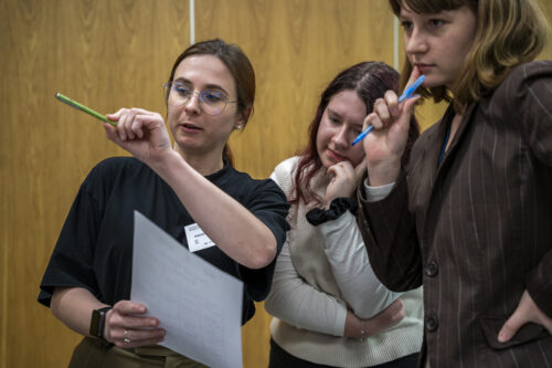 Zbliżenie na 3 młode dziewczyny stojące w pomieszczeniu. Dziewczyny przyglądają się czemuś co jest poza kadrem. Dziewczyna stojąca najbardziej po lewej stronie ma rude włosy. Ma też wyciągniętą lewą rękę, w której trzyma zielony długopis. Tłumaczy coś pozostałej dwójce. Dziewczyna w środku pochyla się do przodu. Dłonią podpiera głowę. Trzecia z dziewczyn stoi najbardziej po prawej. Trzyma w ręce niebieski długopis. Przykłada go do ust w zamyśleniu.